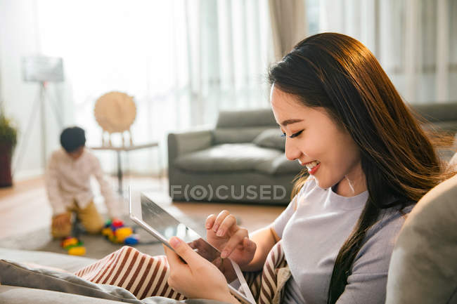 Красива усміхнена молода азіатська жінка використовує цифровий планшет, коли син грає з іграшками позаду вдома — стокове фото