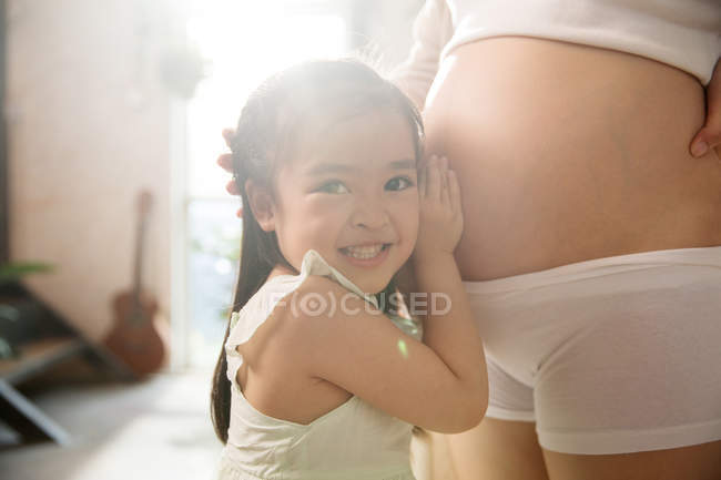 Обрізаний знімок щасливої маленької дівчинки, що слухає живіт вагітної матері — стокове фото