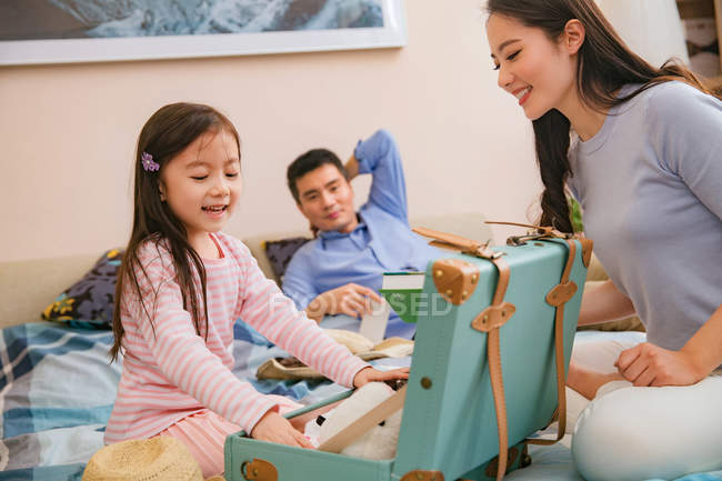 Genitori felici guardando adorabile piccola figlia valigia di imballaggio sul letto — Foto stock