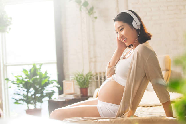 Jovem mulher grávida relaxada sentada na cama e ouvir música em fones de ouvido — Fotografia de Stock