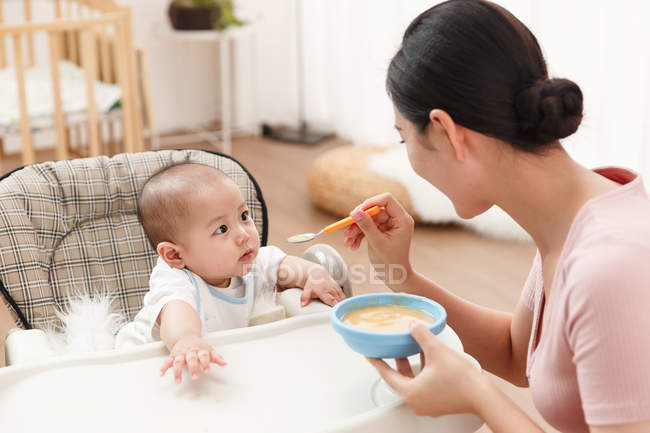 Felice giovane madre che tiene ciotola e cucchiaio, alimentando adorabile bambino a casa — Foto stock