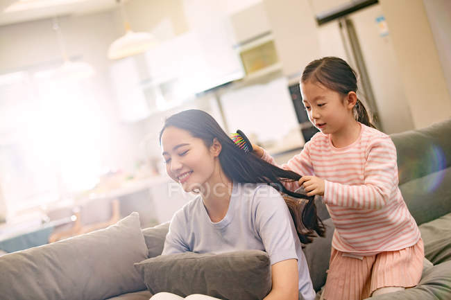 Чарівна маленька дочка розчісує волосся до усміхненої молодої матері, що сидить на дивані — стокове фото