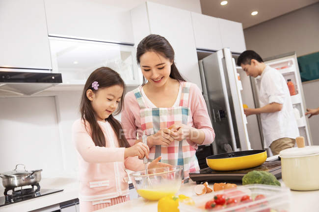 Mãe feliz e filha cozinhar juntos, enquanto o pai abertura geladeira atrás na cozinha — Fotografia de Stock