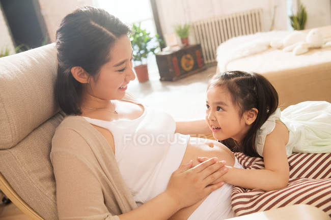 Прелестная счастливая маленькая девочка обнимает и слушает живот беременной матери — стоковое фото