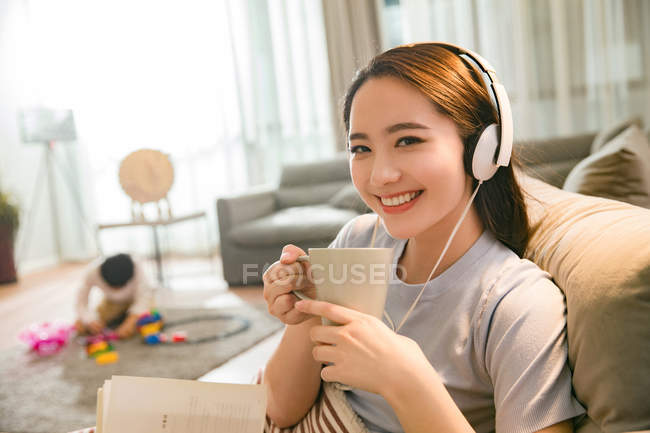 Молода китайка в навушниках тримає чашку і посміхається на камеру, коли син грає з іграшками позаду вдома — стокове фото