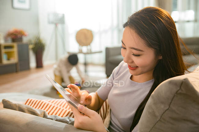 Hermosa sonriente joven asiático mujer usando digital tablet mientras hijo jugando con juguetes detrás en casa - foto de stock