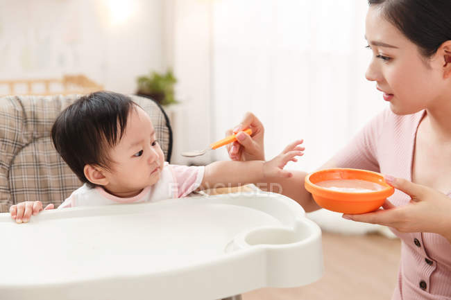 Colpo ritagliato di giovane madre che tiene la ciotola con il cucchiaio e alimenta il bambino adottabile a casa — Foto stock