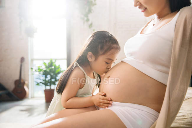 Вид сбоку восхитительной маленькой дочки, целующей животик беременной матери, обрезанный снимок — стоковое фото