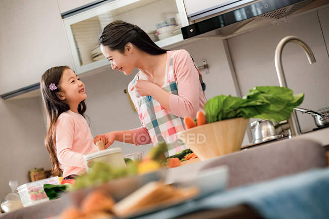 Щаслива азіатська мати і дочка посміхаються один одному, готуючи разом на кухні — стокове фото