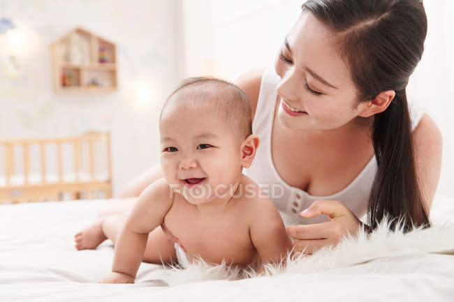 Glückliche junge Mutter schaut entzückendes Baby in Windel lachend an und blickt in die Kamera — Stockfoto