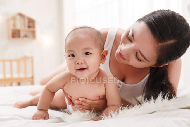Feliz jovem mãe olhando adorável bebê deitado na cama e sorrindo para a câmera — Fotografia de Stock