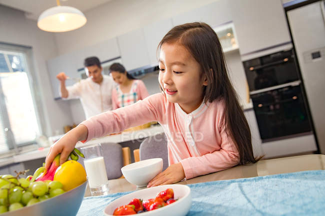 Adorable niño comiendo frutas mientras padres cocinando detrás en cocina - foto de stock