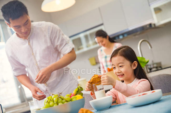 Pai com adorável sorrindo filha à mesa com café da manhã, mãe cozinhar atrás na cozinha — Fotografia de Stock