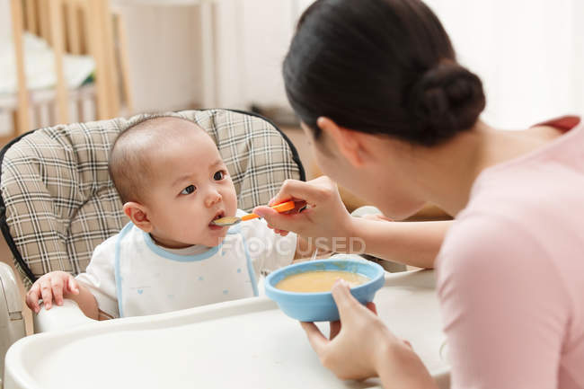 Young asian woman feeding cute baby at home — Stock Photo