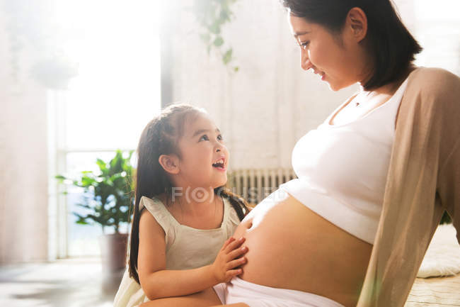 Adorable feliz niña mirando embarazada madre en casa - foto de stock