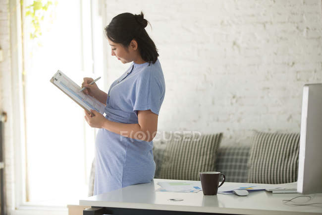 Visão lateral da jovem empresária grávida escrevendo na área de transferência — Fotografia de Stock