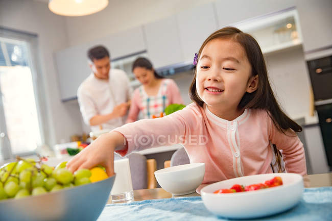 Adorável criança comer frutas enquanto os pais cozinhar atrás na cozinha — Fotografia de Stock