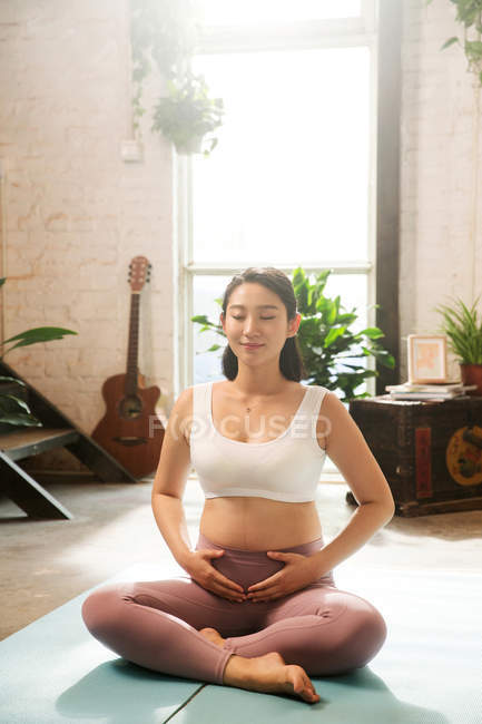 Felice giovane donna incinta in abbigliamento sportivo seduta su tappetini yoga a casa — Foto stock