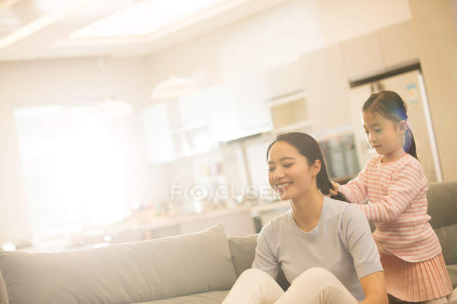 Adorable little daughter plaiting braid to smiling young mother sitting on couch — Stock Photo