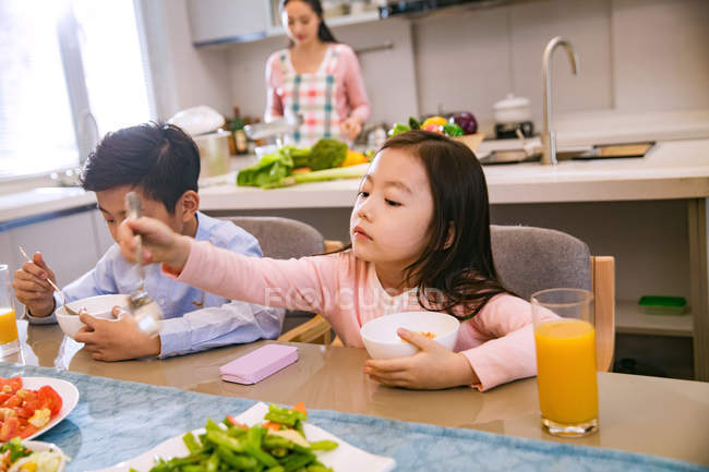 Adorable asian brother and sister having breakfast together while mother cooking behind in kitchen — Stock Photo