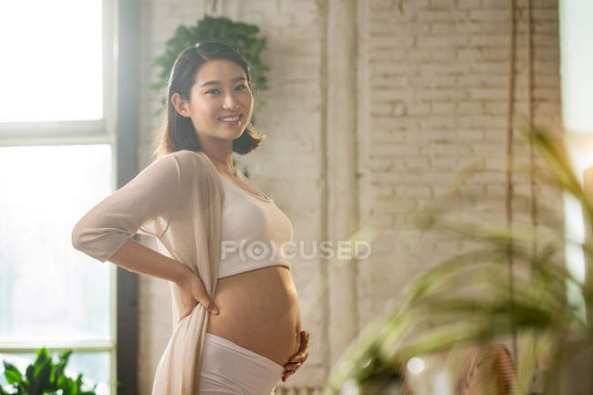 Vista lateral de alegre joven asiática embarazada tocando vientre y sonriendo a la cámara, enfoque selectivo - foto de stock