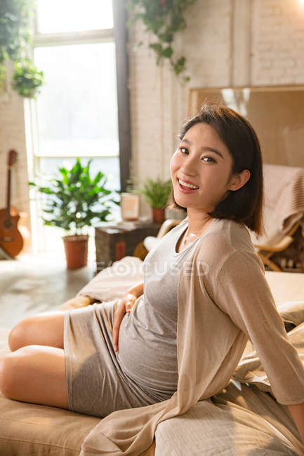 Glückliche junge schwangere Frau sitzt auf dem Bett und lächelt in die Kamera — Stockfoto