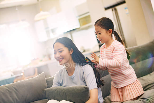 Entzückende kleine Tochter kämmt Haare für lächelnde junge Mutter, die auf der Couch sitzt — Stockfoto