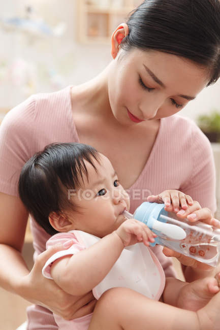 Happy young mother looking at baby drinking from bottle at home — Stock Photo