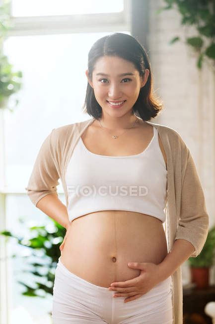 Heureux jeune enceinte asiatique femme toucher ventre et sourire à la caméra — Photo de stock