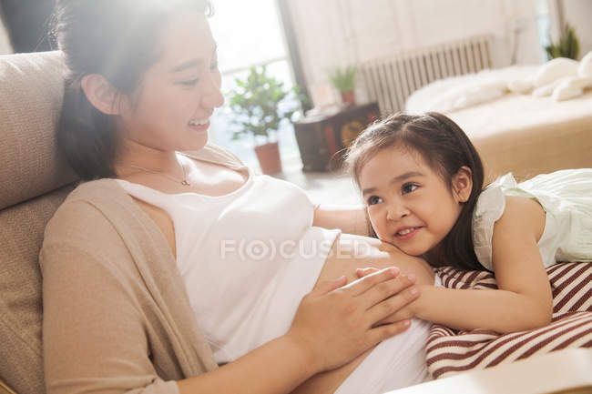Прелестная счастливая маленькая девочка обнимает и слушает живот беременной матери — стоковое фото