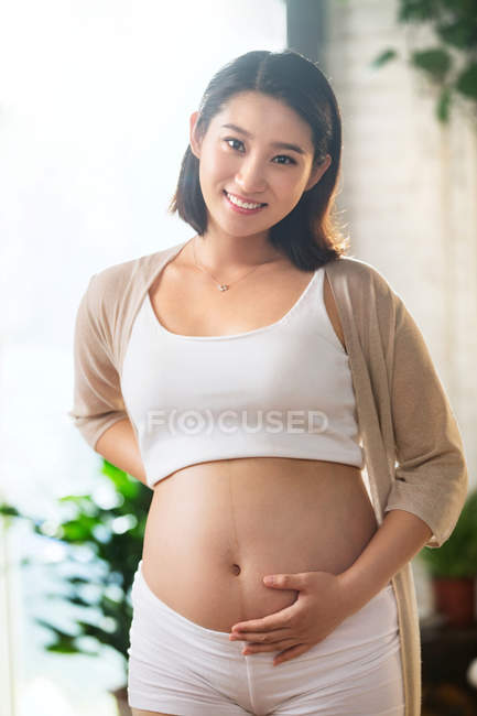 Счастливая молодая беременная азиатка трогает живот и улыбается в камеру — стоковое фото