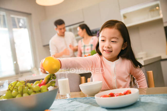 Entzückend lächelndes Kind hält Zitrone in der Hand, während Eltern in der Küche hinterherkochen — Stockfoto