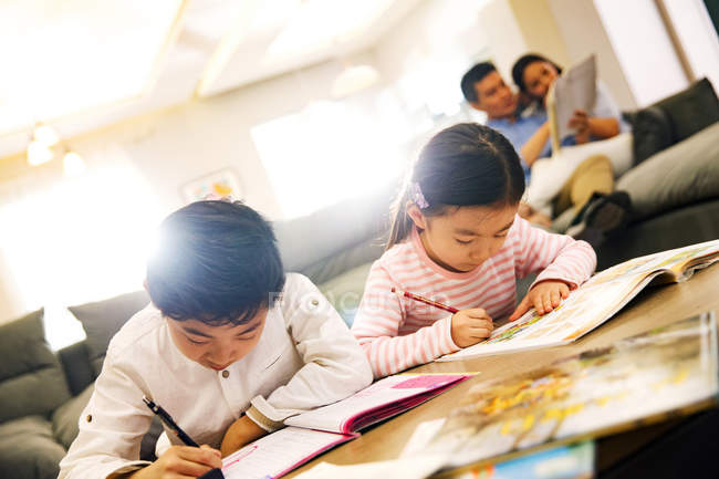 Adorabili bambini cinesi che fanno i compiti a casa, i genitori seduti dietro — Foto stock