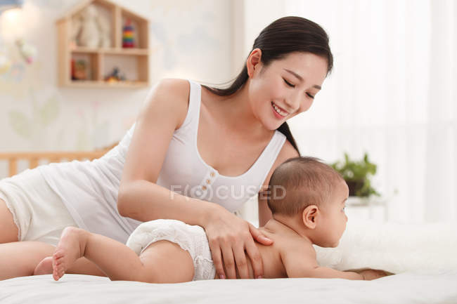 Счастливая молодая мать смотрит на очаровательный ребенок в подгузнике лежа на кровати — стоковое фото