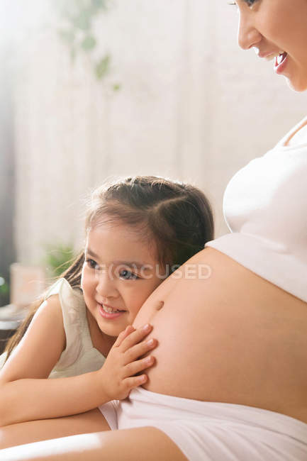 Прелестная счастливая маленькая девочка обнимает и слушает живот беременной матери дома, обрезанный выстрел — стоковое фото
