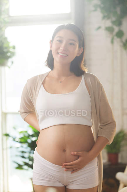 Cheerful young asian pregnant woman touching belly and smiling at camera — Stock Photo