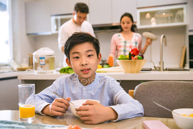 Menino chinês bonito sentado à mesa e olhando para a câmera, pais de pé atrás na cozinha — Fotografia de Stock