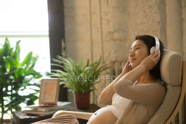 Молодая расслабленная беременная женщина сидит в кресле и слушает музыку в наушниках — стоковое фото