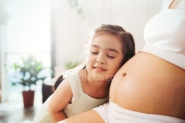 Чарівна щаслива дівчинка обіймає і слухає живіт вагітної матері вдома, обрізаний постріл — стокове фото