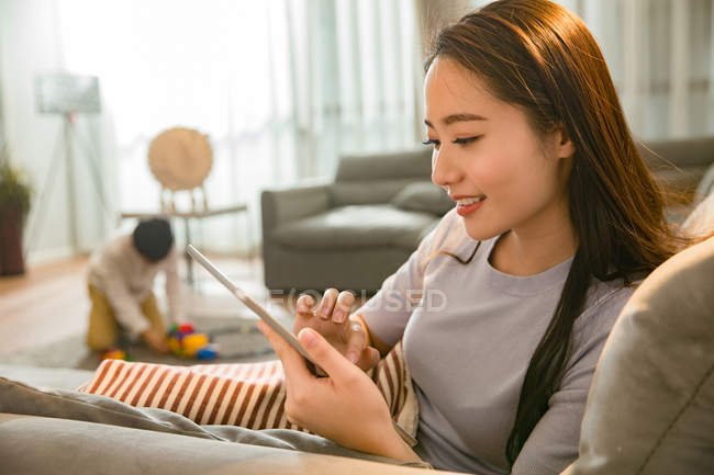 Schöne lächelnde junge Asiatin mit digitalem Tablet, während ihr Sohn zu Hause mit Spielzeug spielt — Stockfoto