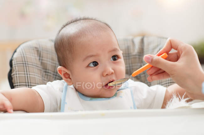 Colpo ritagliato di madre che tiene cucchiaio e alimenta adorabile bambino a casa — Foto stock