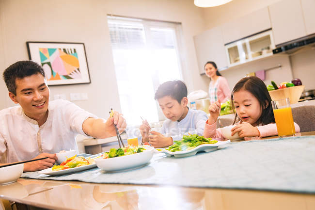 Pai com crianças sentadas e comendo à mesa, mãe cozinhar na cozinha — Fotografia de Stock