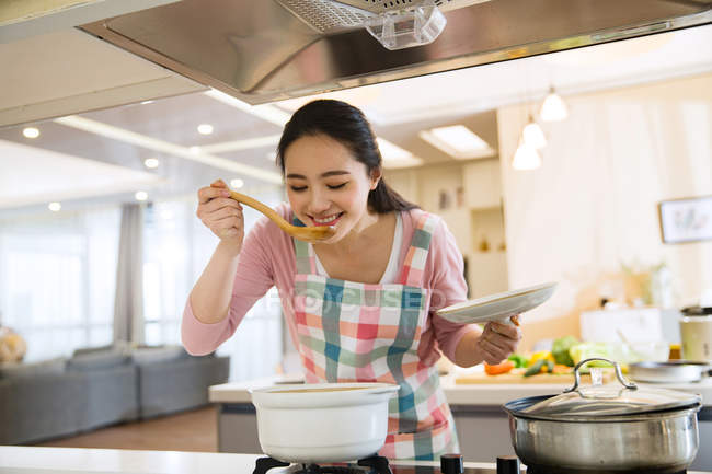 Souriant jeune femme cuisine et plat de dégustation dans la cuisine — Photo de stock