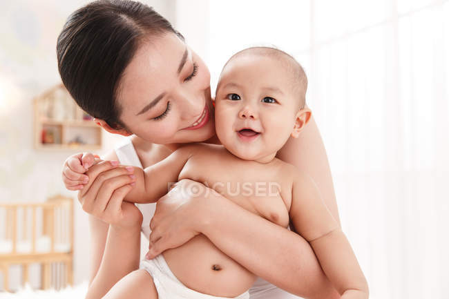 Красива щаслива молода азіатська жінка обіймає чарівну дитину в пелюшці вдома — стокове фото