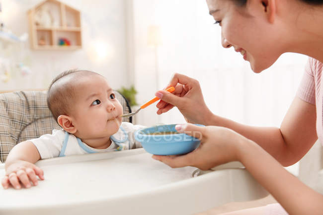 Feliz jovem mãe segurando tigela e colher, alimentando bebê adorável em casa — Fotografia de Stock
