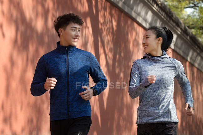 Athletic young couple in sportswear smiling each other and running together on street — Stock Photo