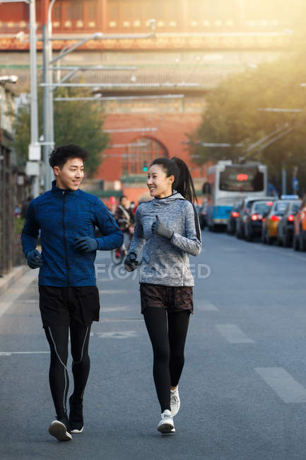 Jóvenes atletas asiáticos en ropa deportiva sonriendo unos a otros y trotando juntos en la calle - foto de stock