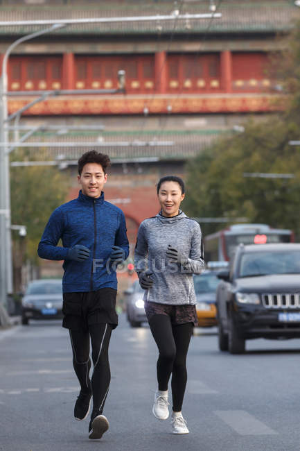 Счастливая спортивная молодая азиатская пара, улыбающаяся в камеру и бегающая вместе по улице — стоковое фото