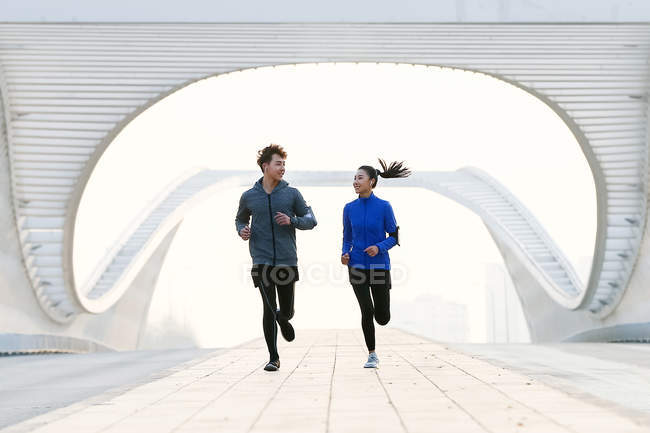 Vista frontal de la feliz joven pareja asiática en ropa deportiva sonriendo entre sí y corriendo juntos en el puente - foto de stock