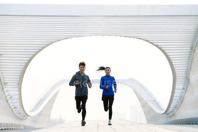 Vista frontal de los jóvenes atletas sonrientes masculinos y femeninos corriendo juntos en el puente moderno - foto de stock